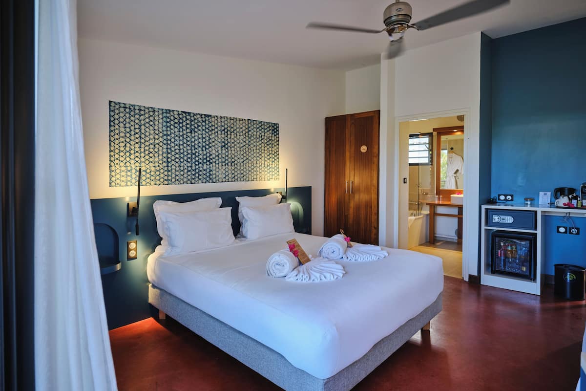 Quarto de hotel com cama, ventoinha no teto, banheiro visível por uma porta aberta e minibar.