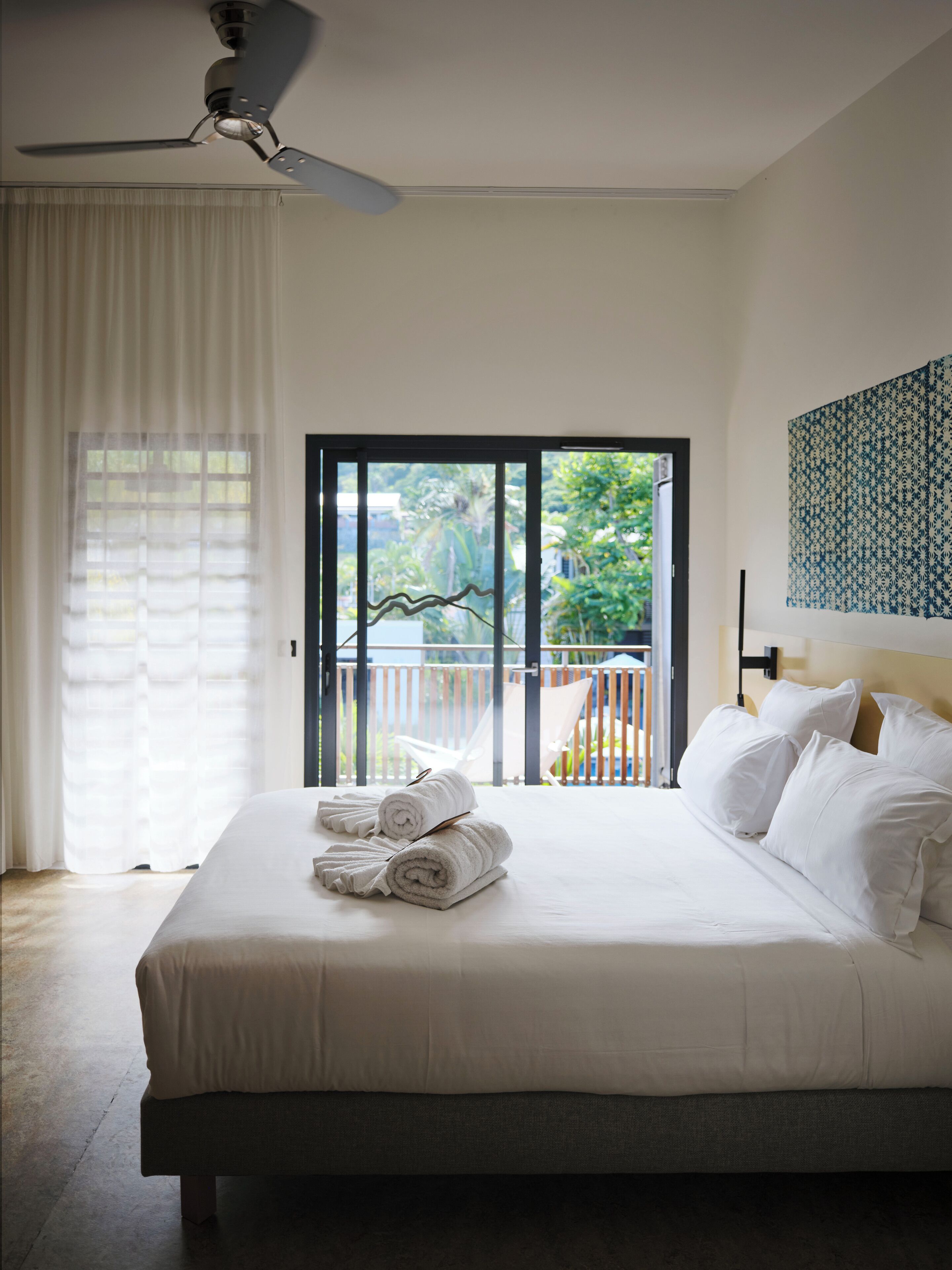 A hotel room with a bed, white towels, a ceiling fan, a view of greenery through a sliding door, and a patterned wall hanging.