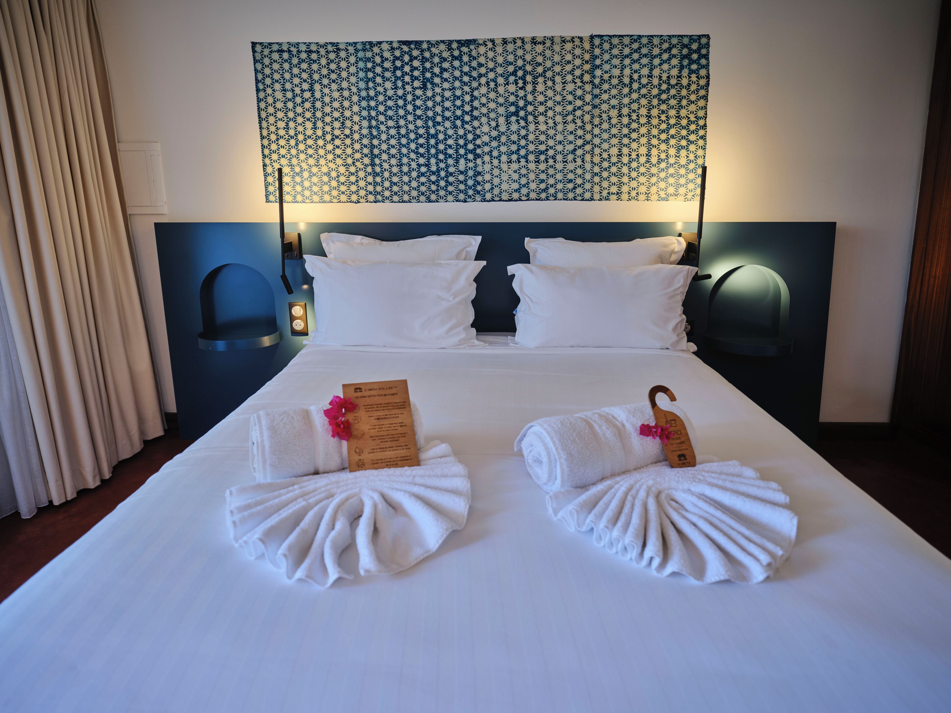 A hotel room with a neatly made bed, two towels with a card, and a patterned headboard.