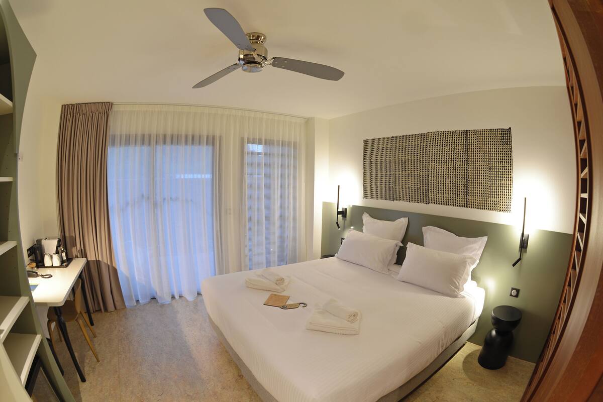 Quarto de hotel com cama, escrivaninha com cadeira, ventilador de teto e janela com cortinas.