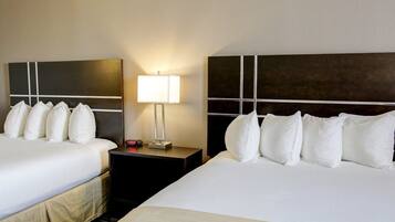 Habitación de hotel con dos camas, cada una con sábanas blancas y varias almohadas. Hay una mesita de noche con lámpara y reloj entre las camas.