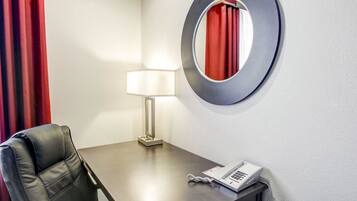 Ett hotellrum med ett skrivbord, en stol, en lampa, en telefon och en rund spegel på väggen.