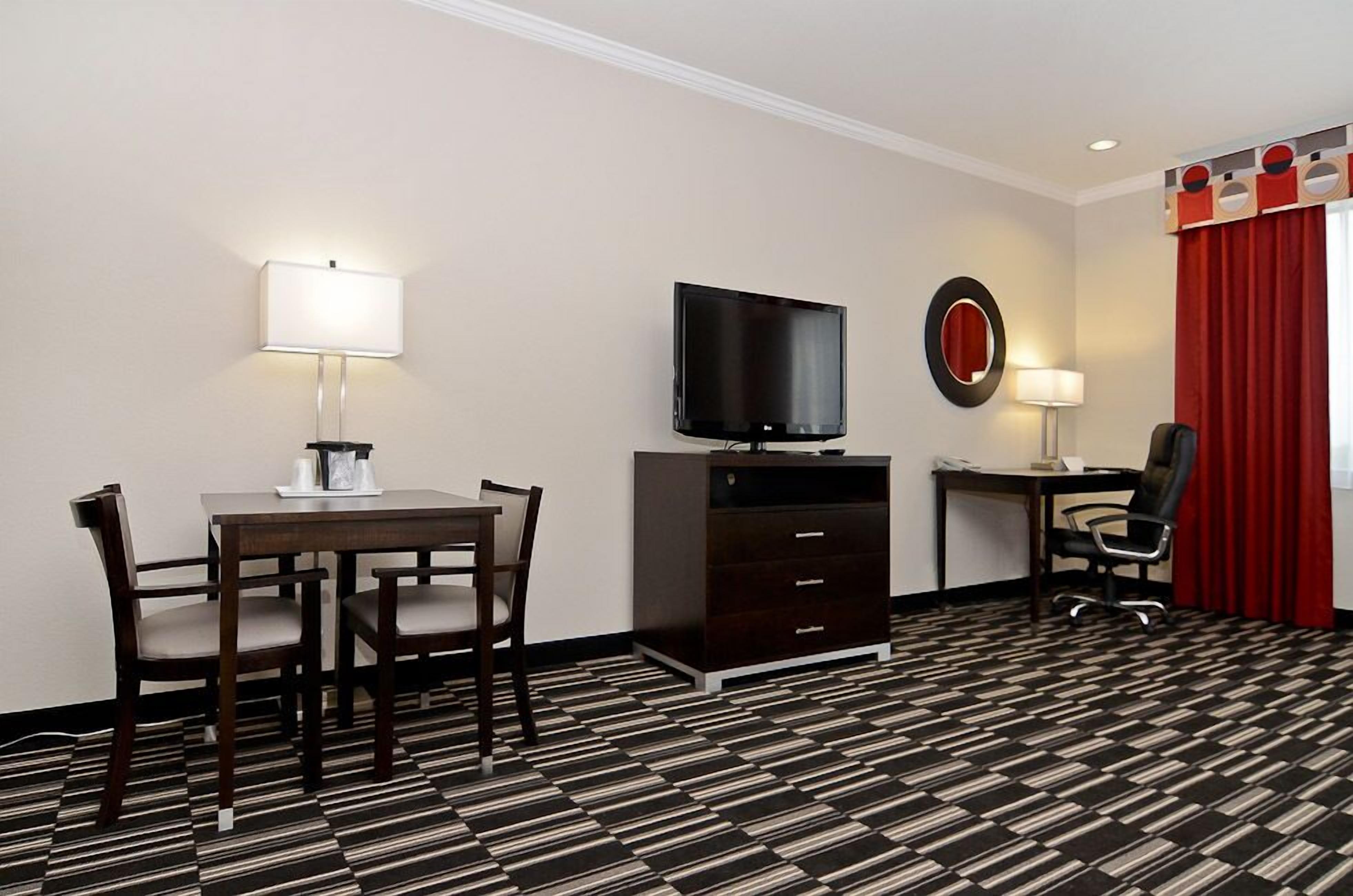 A hotel room with a dining table, chairs, a TV, a desk, a chair, a mirror, and a red curtain.