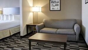 A hotel room with a bed, a gray sofa, a small table, a lamp, and a framed picture on the wall.
