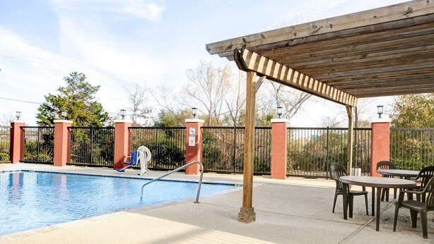 A swimming pool with a wooden pergola and chairs, surrounded by a fenced area with a view of trees.