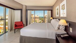 A hotel room with a large bed, a red chair, a desk with a telephone, and a view of buildings and the sea through large windows.