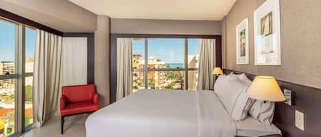 A hotel room with a large bed, a red chair, a desk with a telephone, and a view of buildings and the sea through large windows.