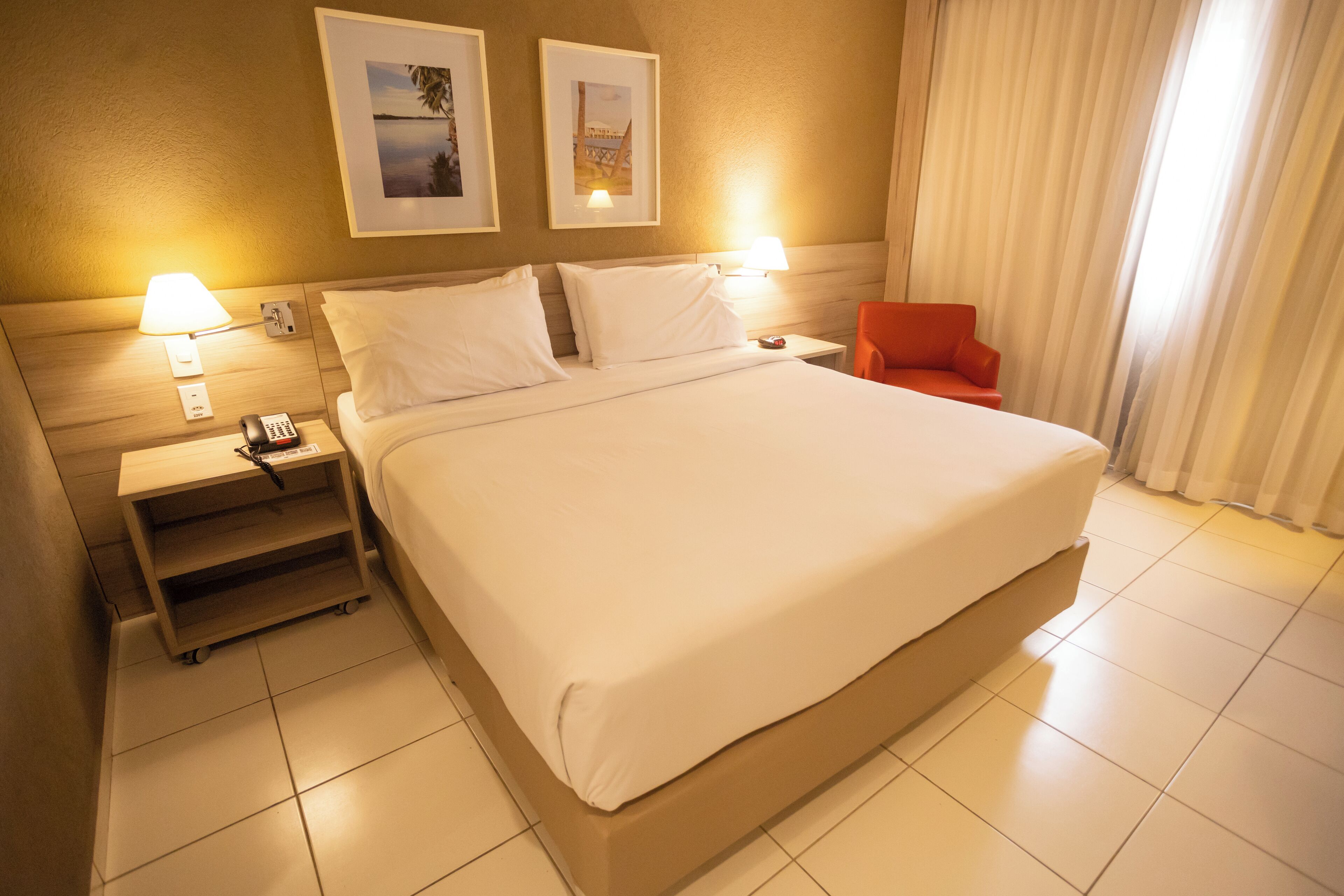 A hotel room with a large bed, two bedside lamps, a nightstand with a phone, a small table, and a red chair.