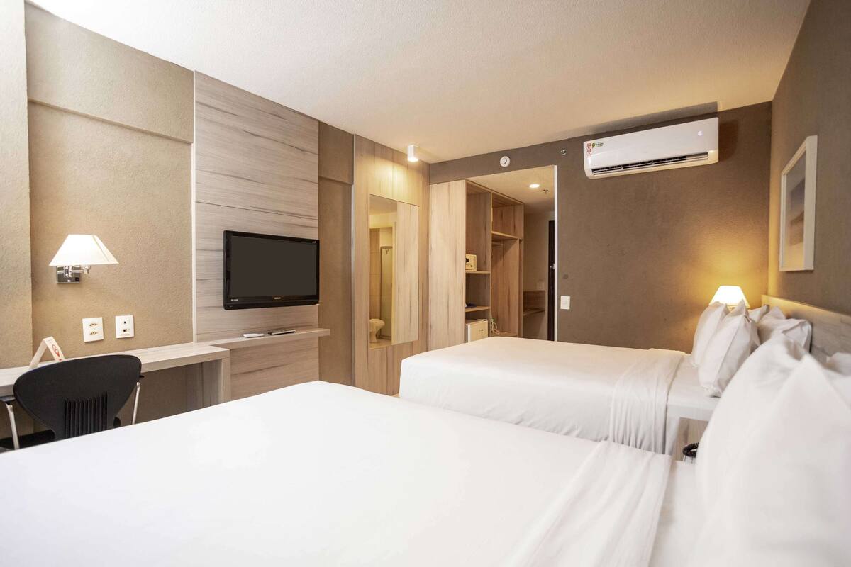 A hotel room with two beds, a desk with a TV, a lamp, an air conditioner, and a wardrobe.
