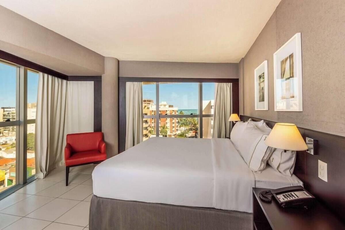 A hotel room with a bed, a red chair, a nightstand with a lamp, and a view of buildings and a body of water.