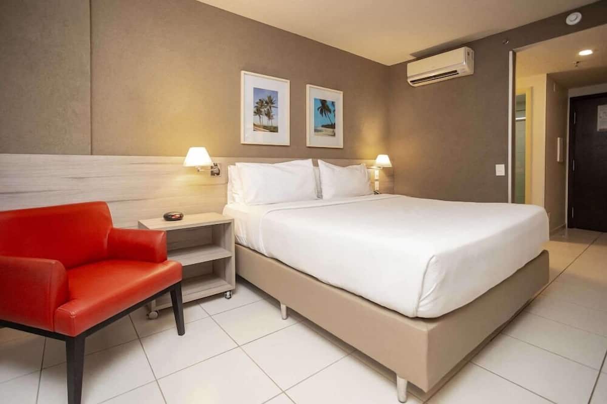 A hotel room with a large bed, two bedside lamps, a red chair, a small table, and two framed pictures on the wall.
