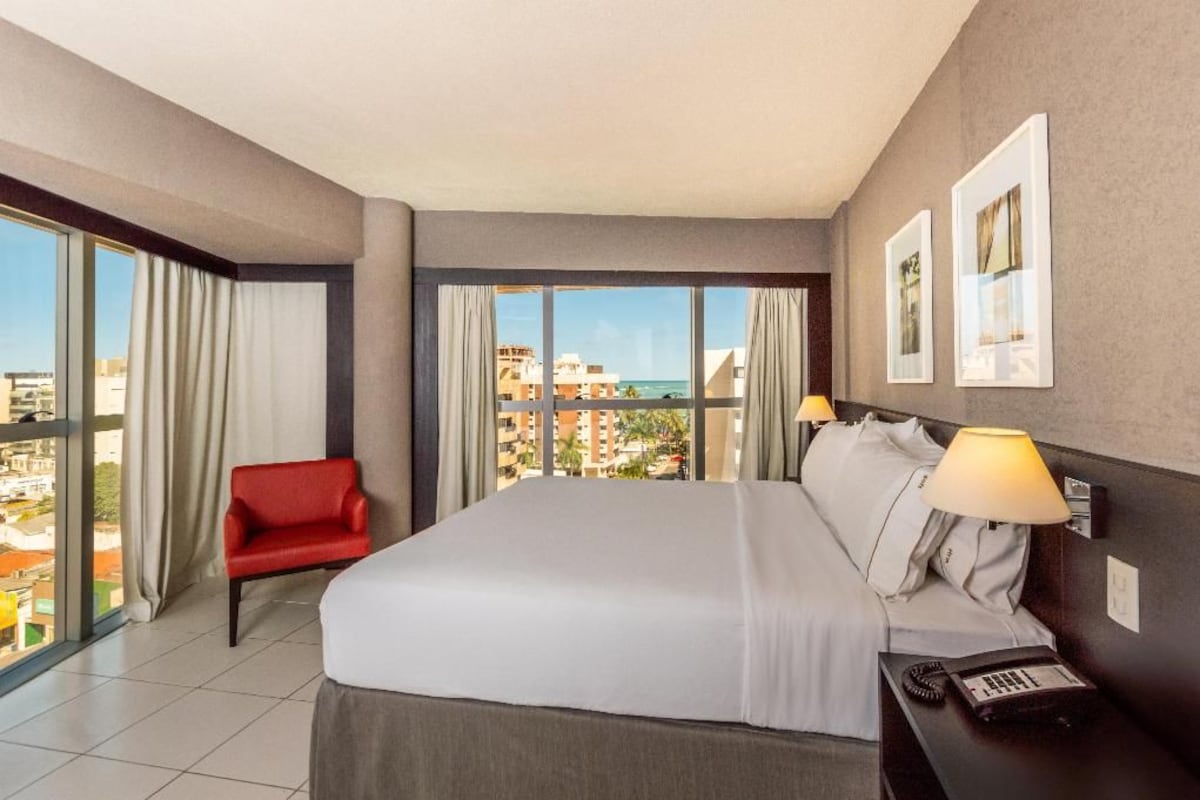 A hotel room with a bed, a red chair, a desk with a lamp, and a view of buildings and water.