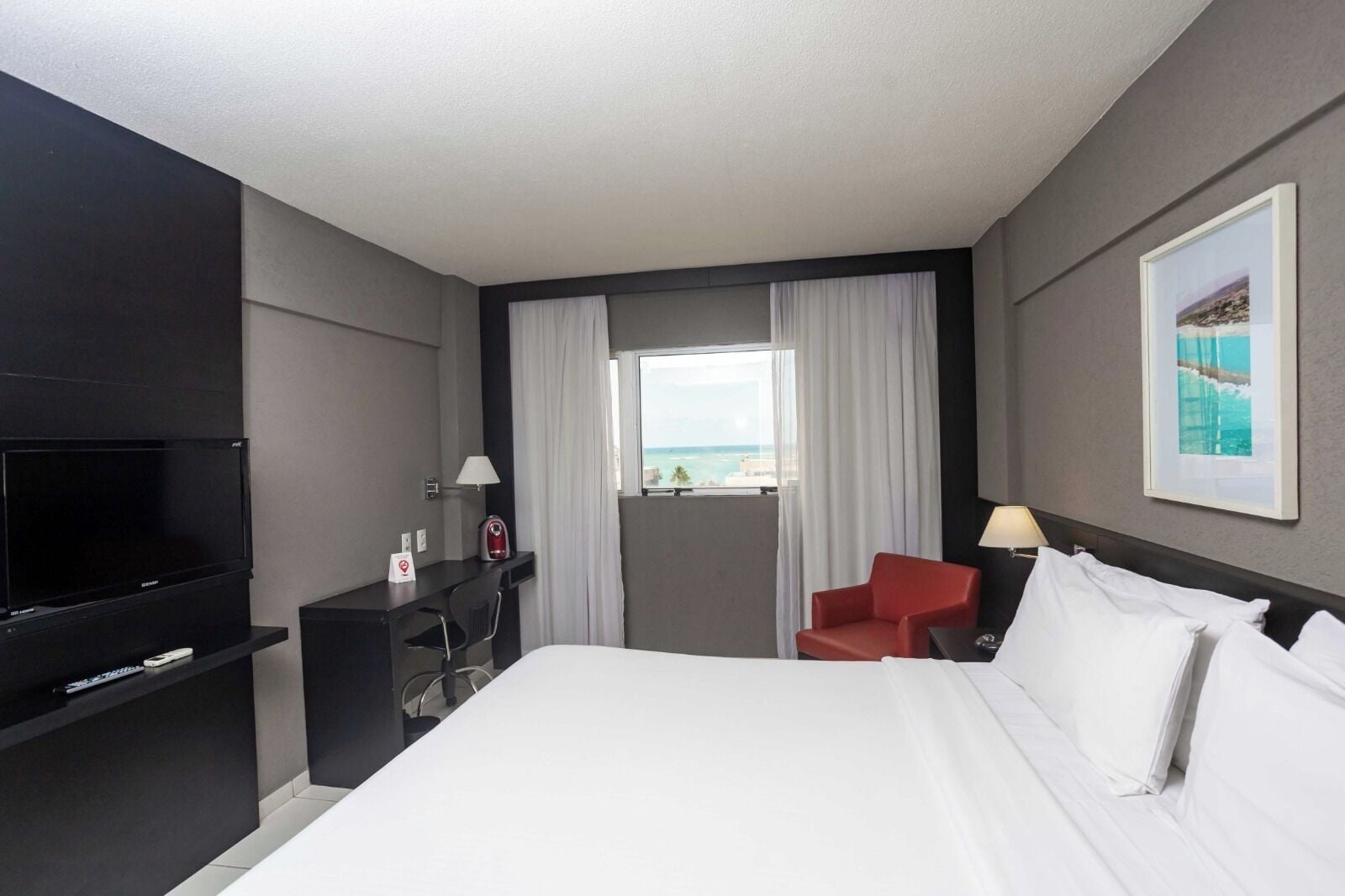 Ein Hotelzimmer mit einem großen Bett, einem Schreibtisch mit Fernseher, einem roten Sessel und einem Fenster mit Meerblick.