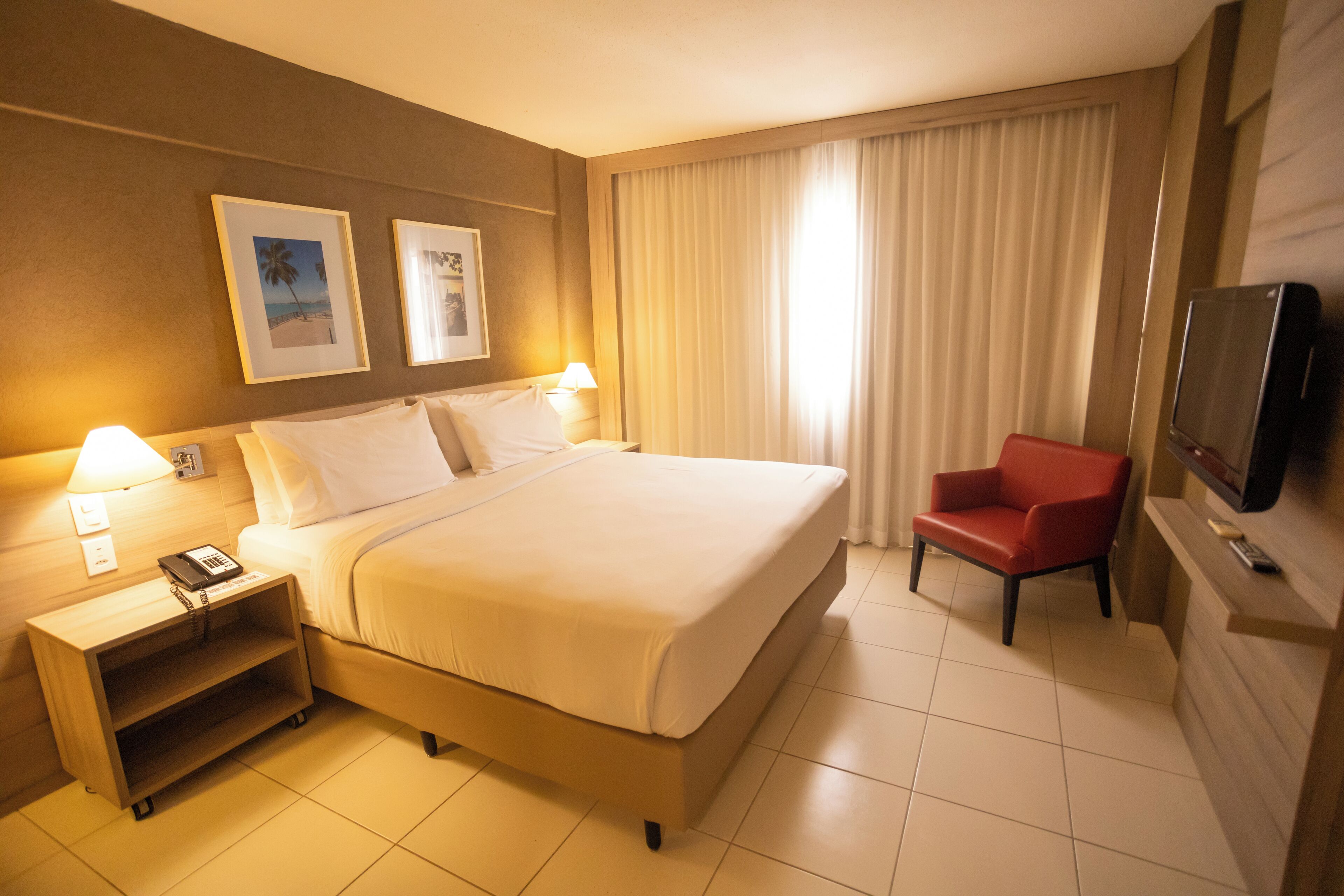 A hotel room with a large bed, a nightstand with a phone, a red chair, a TV, and two framed pictures on the wall.