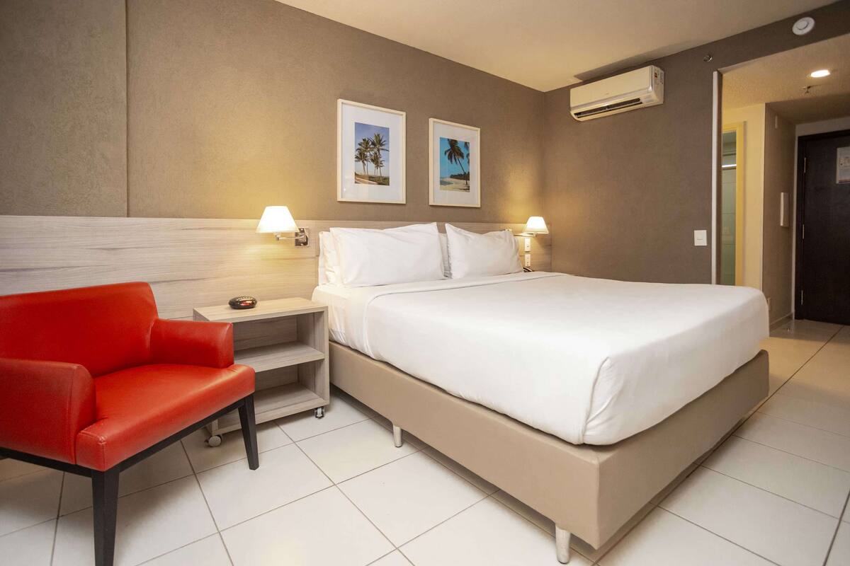 A hotel room with a large bed, two bedside lamps, a red chair, and two framed pictures on the wall.