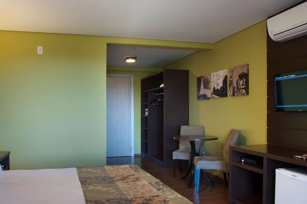 A hotel room with a bed, a desk with a chair, a small table, and a TV. There is a closet and a door leading to another room.
