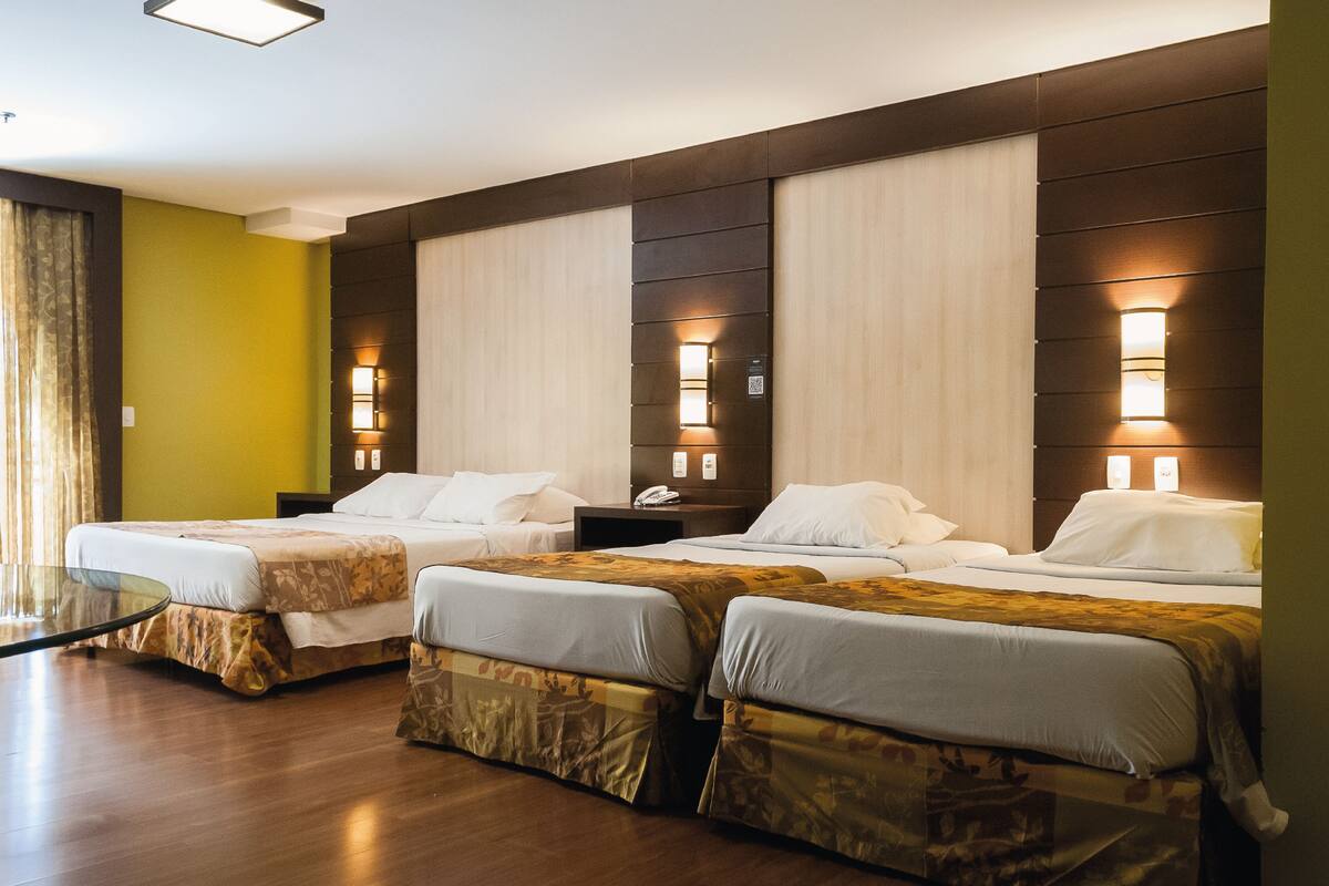 A hotel room with three beds, a glass table, and wall-mounted lamps.