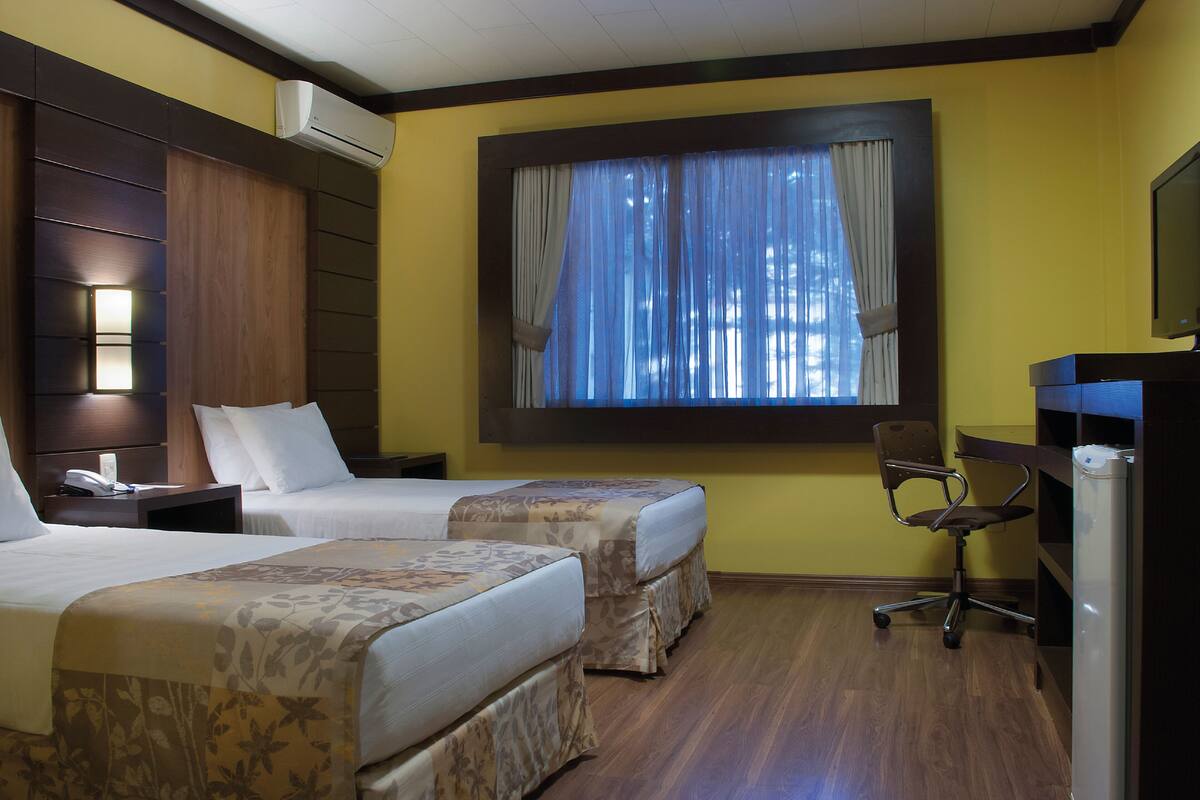 A hotel room with two beds, a desk, a chair, an air conditioning unit, and a window with curtains.
