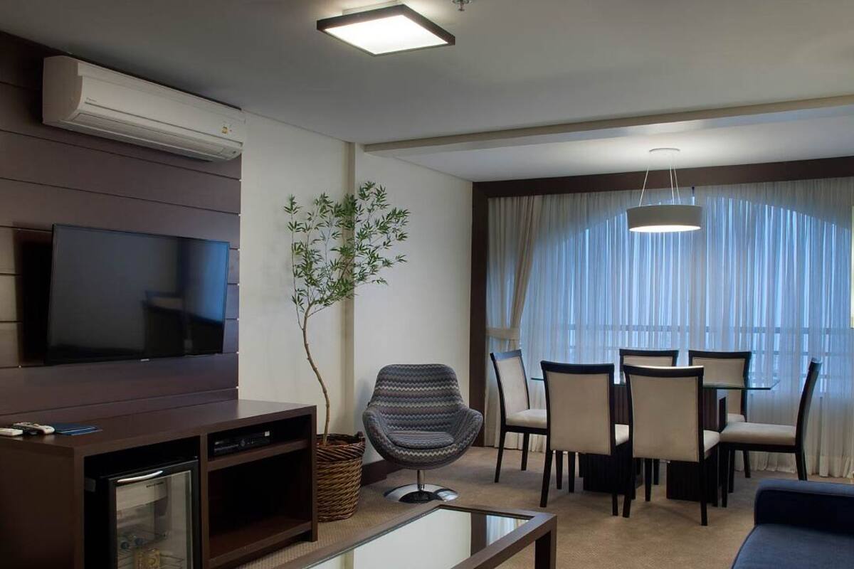 A modern living room with a flat-screen TV, a dining area with a table and chairs, and a large window with curtains.