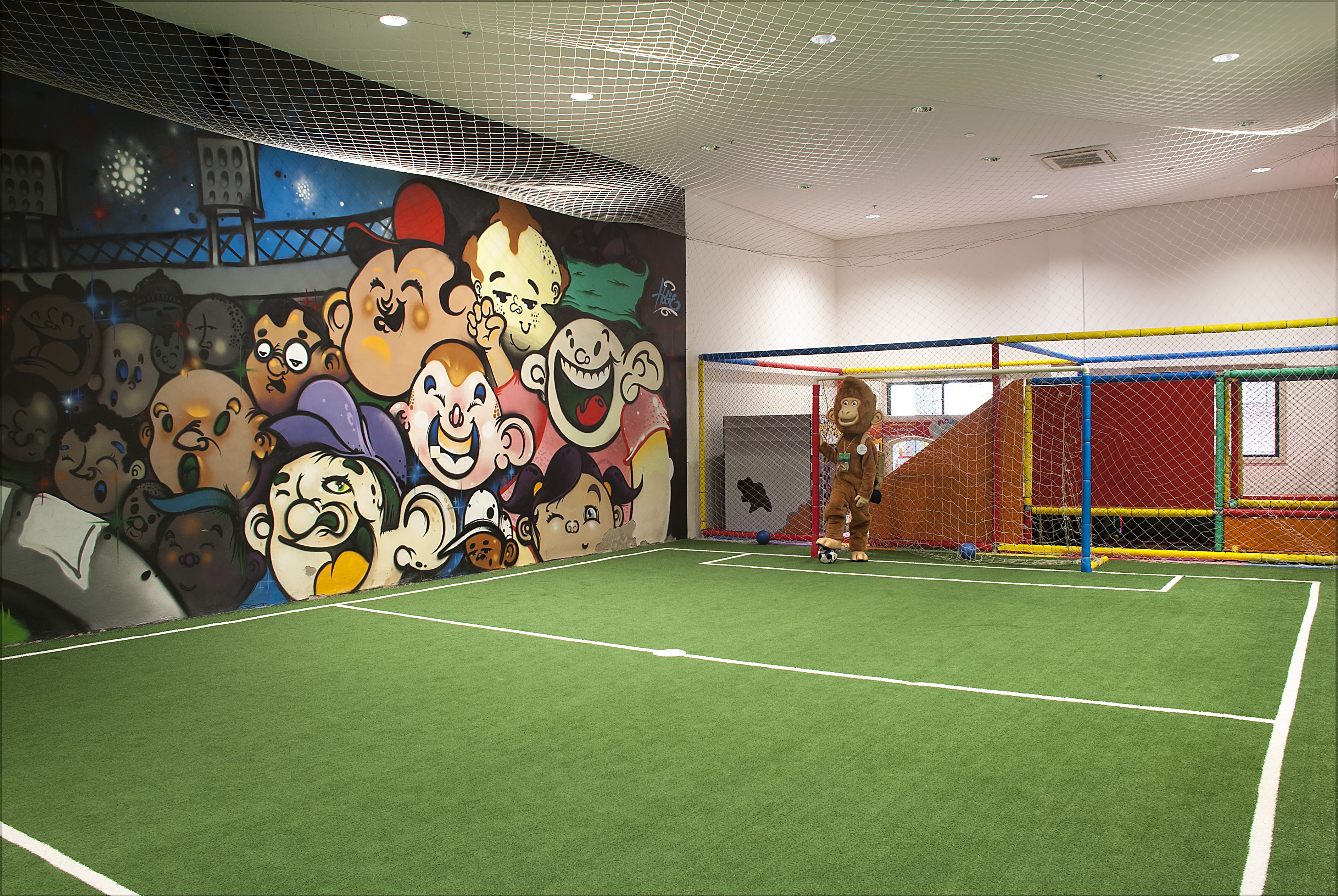 Instalación deportiva interior con campo de fútbol, pared de escalada y un mural de personajes de caricatura.