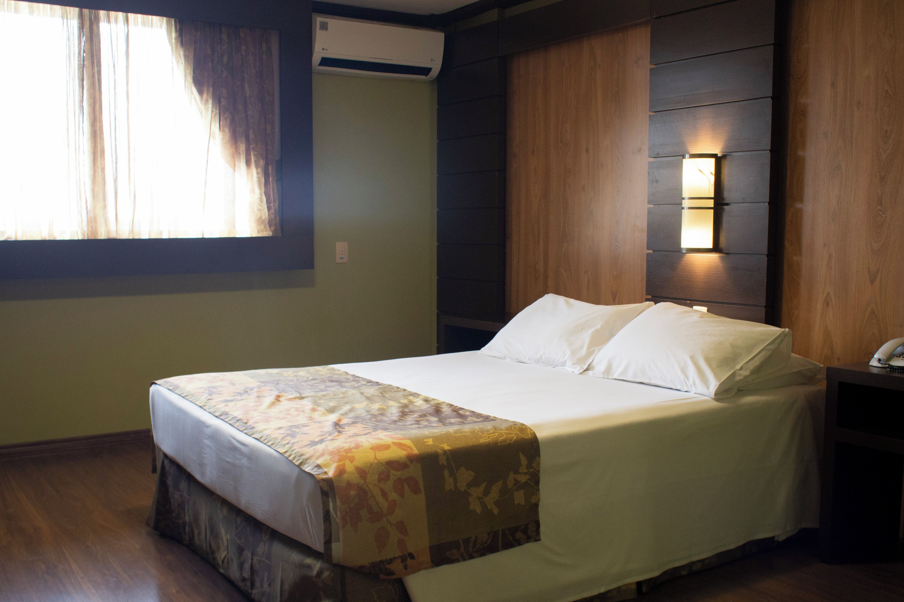 A neatly made bed with a floral bedspread, a wall-mounted light fixture, and a window with curtains.