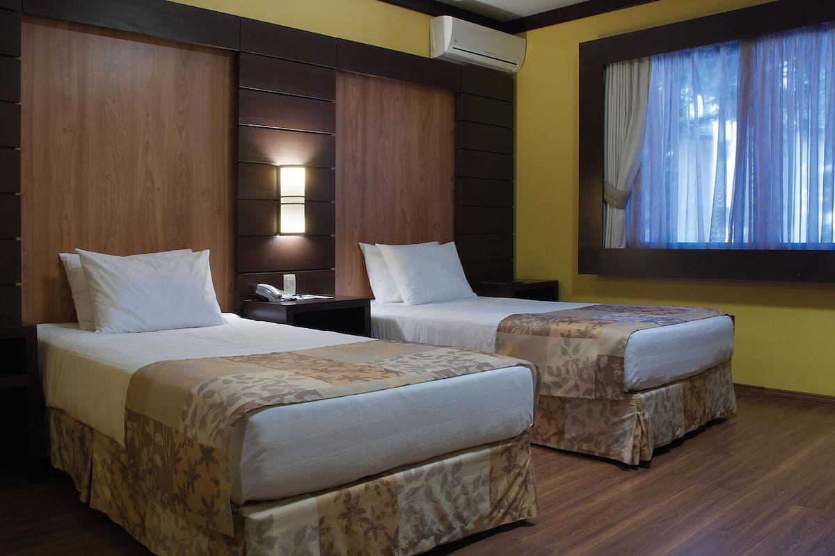 A hotel room with two beds, a nightstand, an air conditioning unit, and a window with curtains.