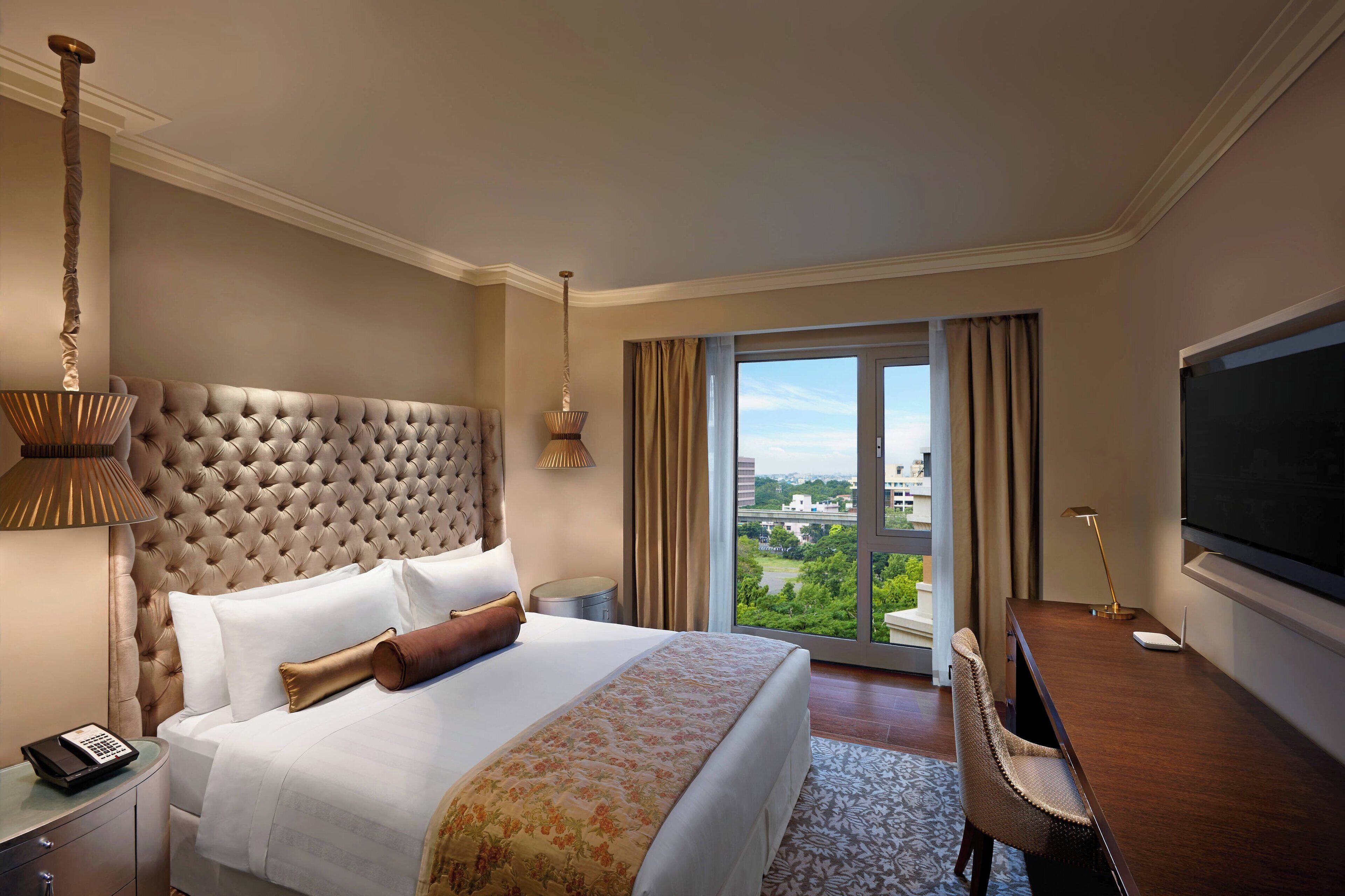 Ein Hotelzimmer mit einer gepolsterten Kopfteil, einem großen Bett mit weißer Bettwäsche, einem Schreibtisch mit Stuhl, einem Flachbildfernseher und Blick ins Grüne durch das Fenster.
