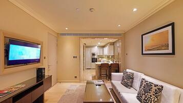 A modern living room with a flat-screen TV, a sofa with patterned cushions, a coffee table, and a kitchen visible through an open doorway.