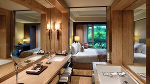 Habitación de hotel con una cama grande, una zona de estar con una silla y una mesa, un espejo amplio, un baño con bañera y vistas a zonas verdes.
