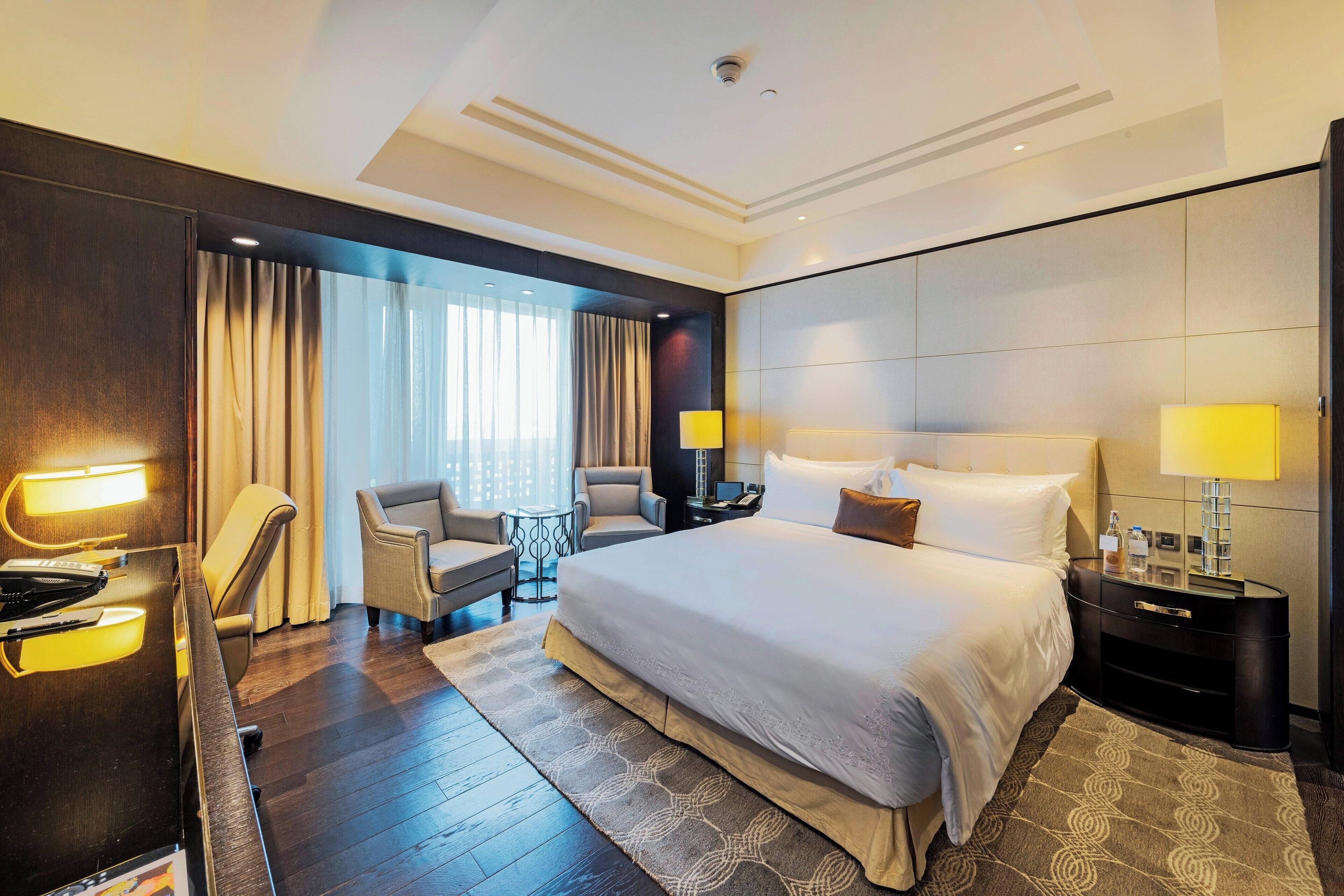A modern hotel room with a large bed, two bedside tables with lamps, a desk with a chair, a small table, and a large window with curtains.