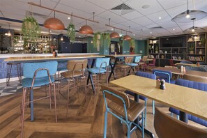 Ein modernes Café mit Holztischen und blauen Stühlen, einer Bar und hängenden Pflanzen.