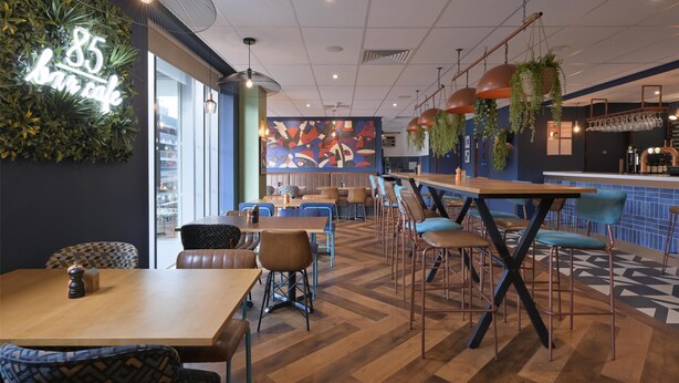A modern cafe interior with wooden tables and chairs, a neon sign, and a colorful mural on the wall.