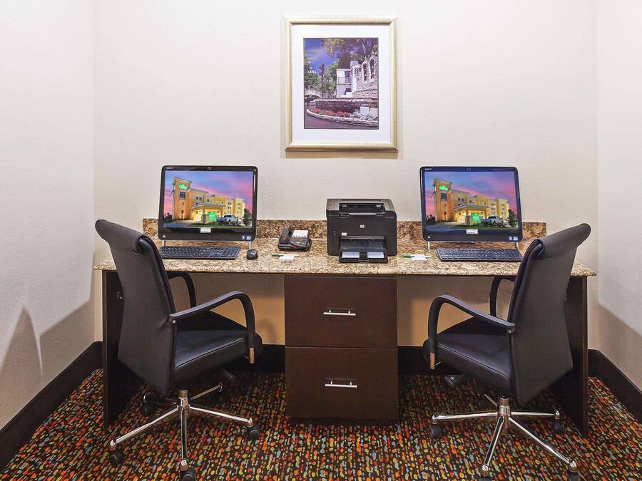 A hotel room with two desks, each with a laptop, a printer, and a telephone. There are two chairs, a framed picture on the wall, and a patterned carpet.