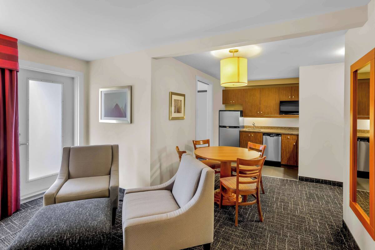 A hotel room with a dining area, a sofa, a small table, and a kitchen area with wooden cabinets and appliances.