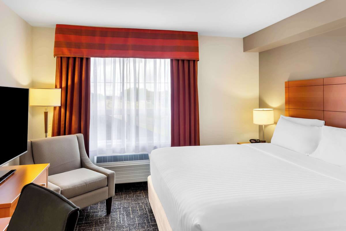 A hotel room with a bed, a desk with a chair, a TV, a window with curtains, and a wall-mounted heater.
