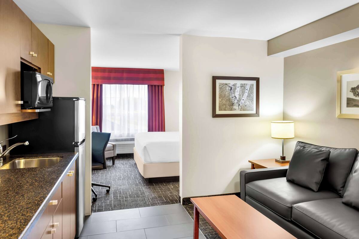 A hotel room with a kitchenette, a bed, a sofa, a desk, and a lamp.