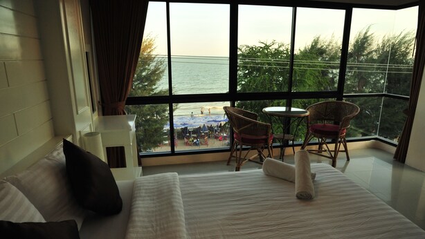 A hotel room with a large window offering a view of a beach and a balcony with wicker chairs and a table. - Magic House Hostel (Cha-am)