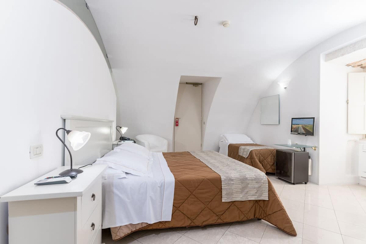 A modern hotel room with a large bed, a desk, a chair, a TV, and a small kitchenette.
