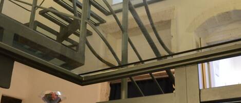 A spiral staircase with a metal frame and glass railings, leading to an upper floor. A wooden table with a decorative glass top is visible on the lower floor.