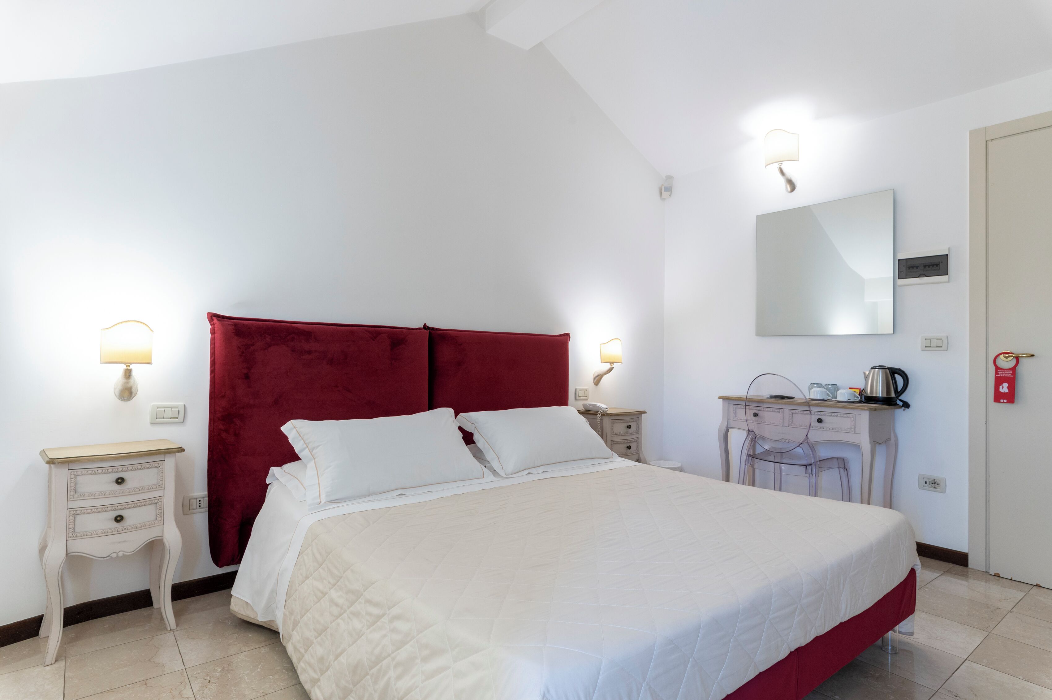 A neatly arranged bedroom with a large bed, a red headboard, bedside tables, a desk with a chair, a mirror, and a wall-mounted light fixture.