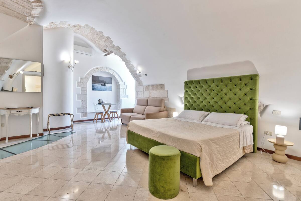 A modern hotel room with a large bed, a green ottoman, a desk, and a chair.