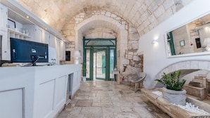 A stone-walled hallway with a modern kitchen area on the left, a seating area with a plant on the right, and an arched doorway leading to another room. - Hotel La Terra (Ostuni)