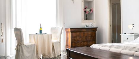 Habitación de hotel con cama, cómoda de madera, una mesita con una botella, dos sillas, un espejo y una ventana con cortinas traslúcidas.