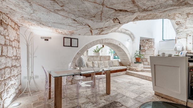 A stone-walled room with a glass table, clear chairs, a sofa, and a kitchen area. - Hotel La Terra (Ostuni)