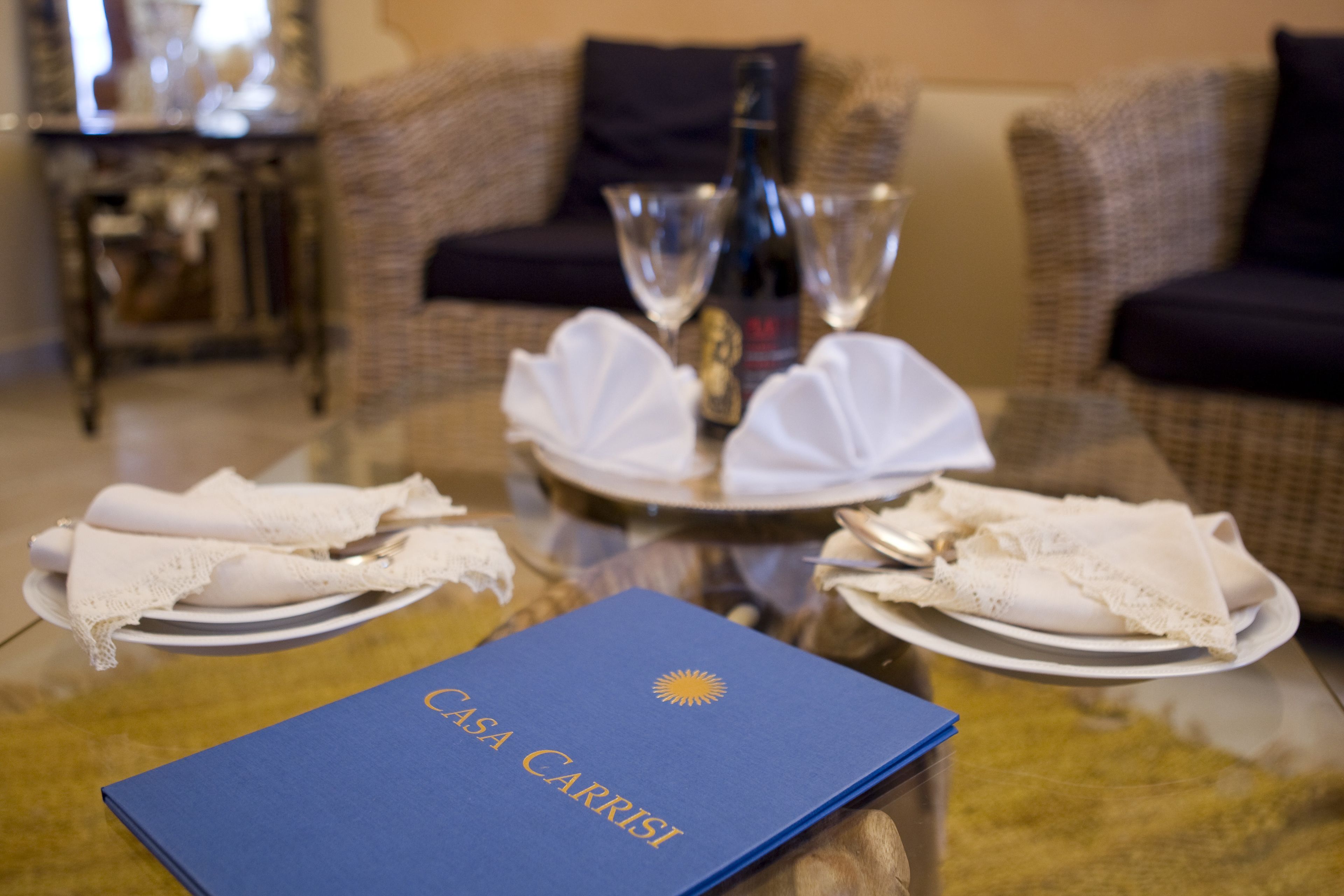 A dining table with a blue folder labeled "Casa Carrisi," two wine glasses, and white napkins on plates.