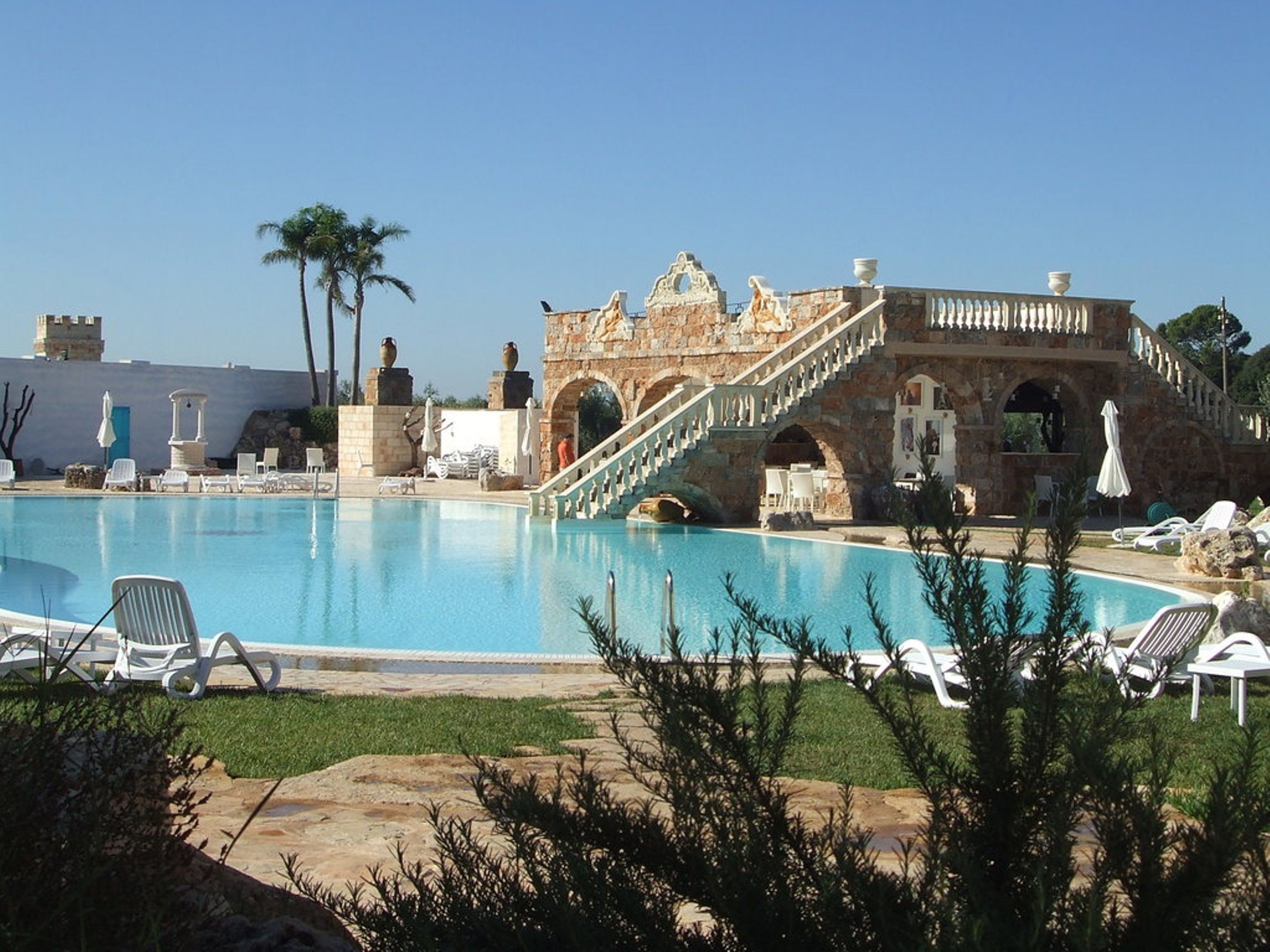 Una piscina con sillas de descanso, un puente de piedra y palmeras.