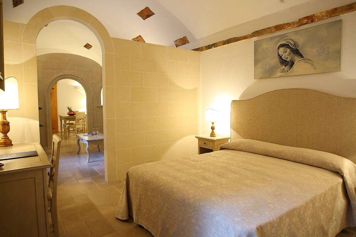 A hotel room with a bed, a desk, and a chair. There is a painting on the wall and a lamp on the desk.