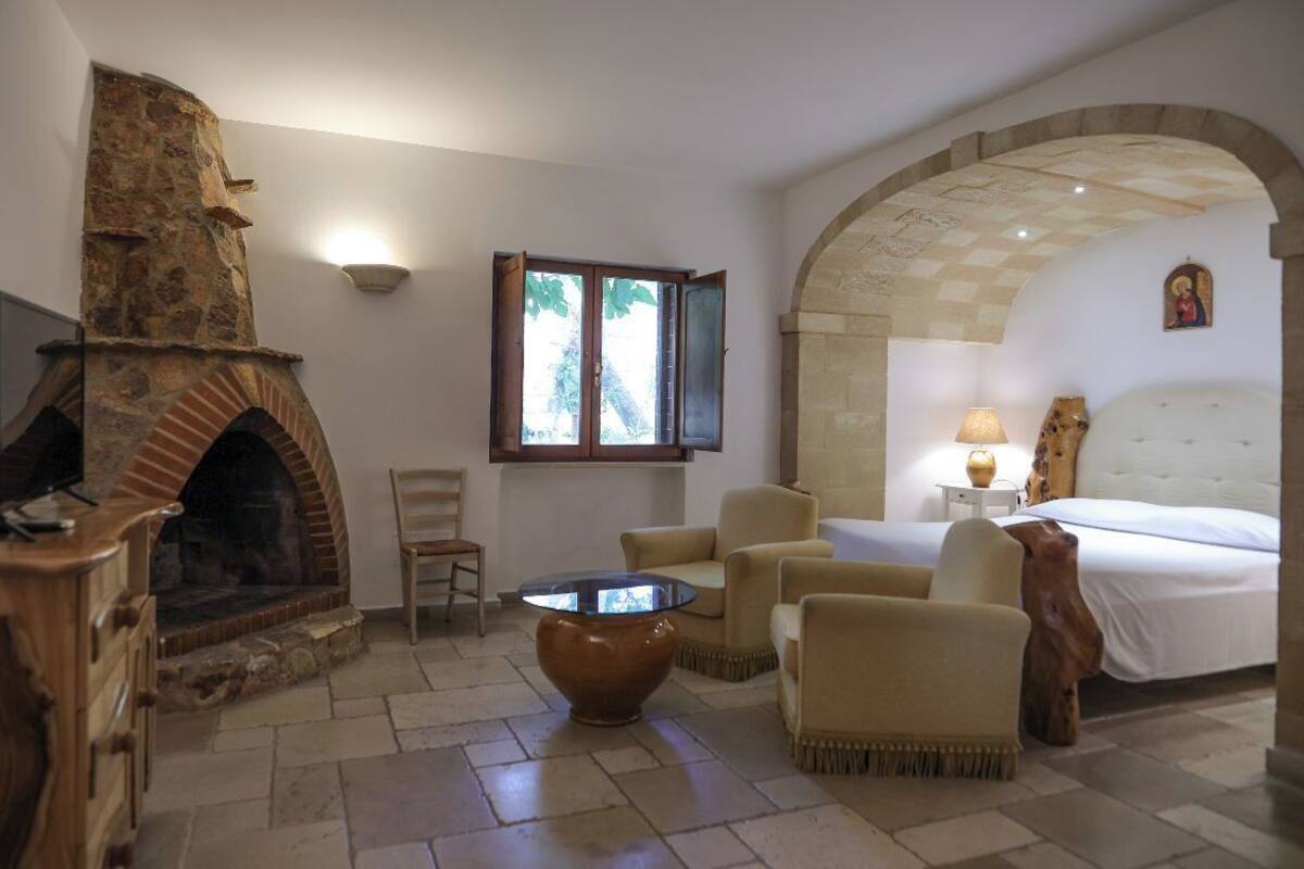 A room with a stone fireplace, a bed, a sitting area with chairs, and a window with a view of trees.