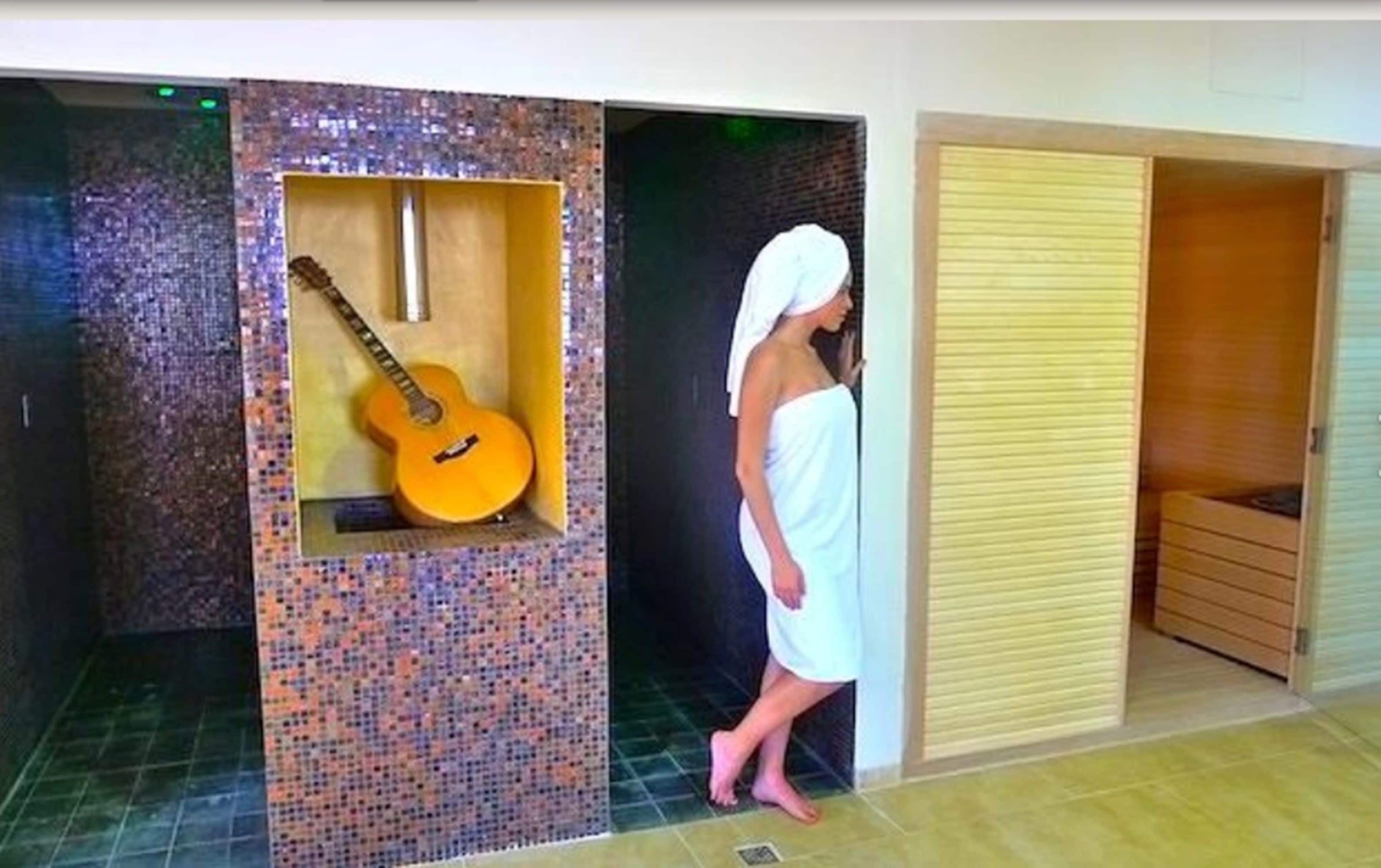 Eine Person in einem weißen Handtuch steht in einem Raum mit einer Wand aus Mosaiken und einer Holztür.