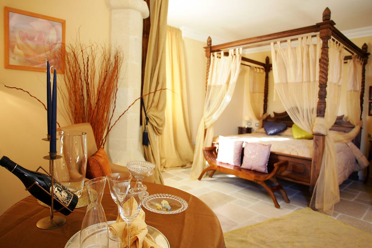 A bedroom with a four-poster bed, a bedside table with a lamp, and a nightstand with a bottle and glasses.